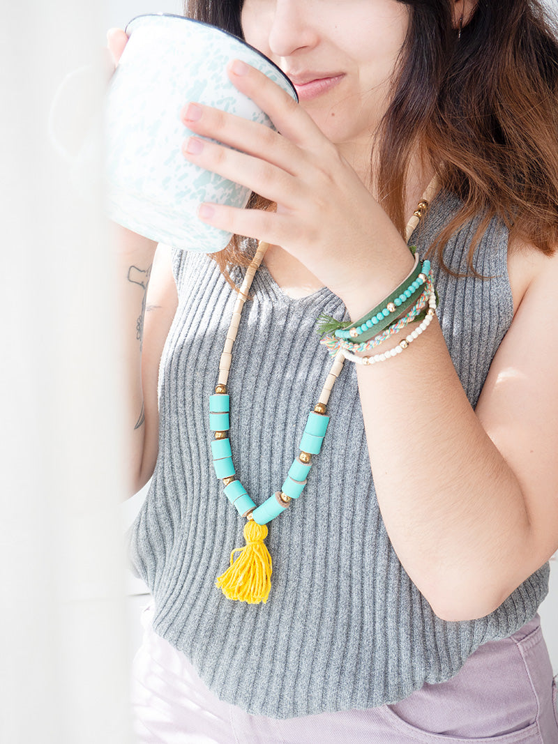 Mujer sonriente con blusa gris usando el collar BOHO en el cuello, en colores turquesa y amarillo