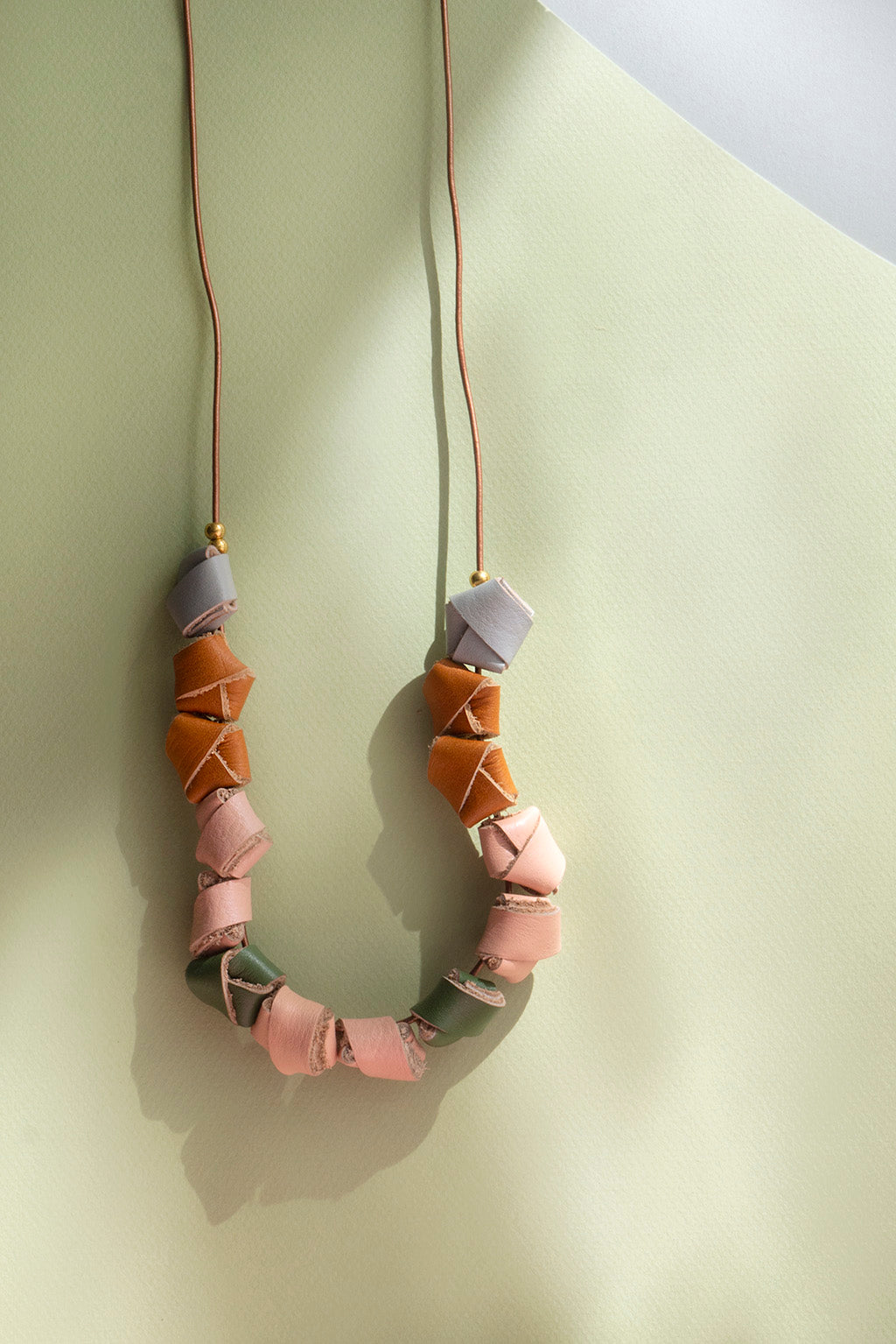 Collar de colores gris, miel, salmón y verde olivo con bolitas doradas y cordón café guindando sobre fondo verde claro